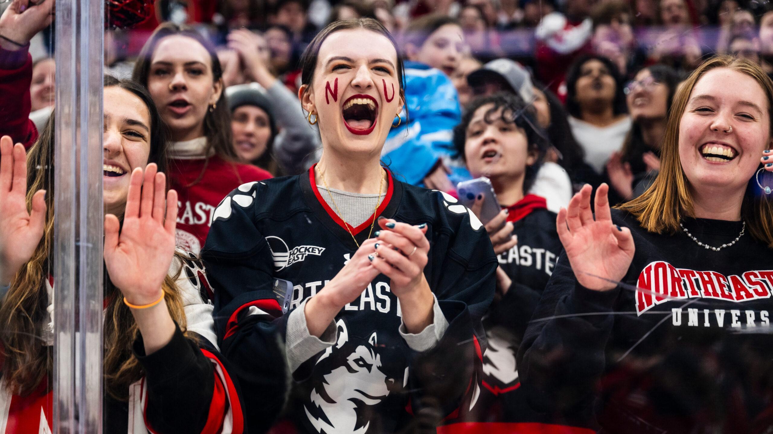 A group of excited Northeastern University fans, wearing team jerseys and sweatshirts, cheer and clap enthusiastically at a hockey game. One fan has "NU" painted on their face. The crowd is lively and energetic.