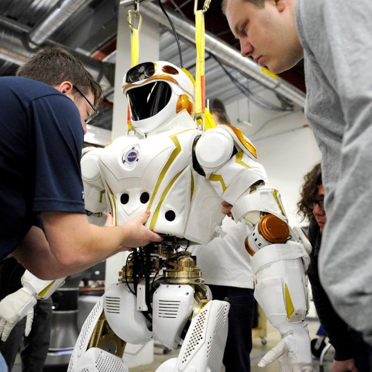 Several people work closely on a suspended humanoid robot with NASA branding in a lab, adjusting its midsection while surrounded by industrial equipment and exposed ductwork overhead.