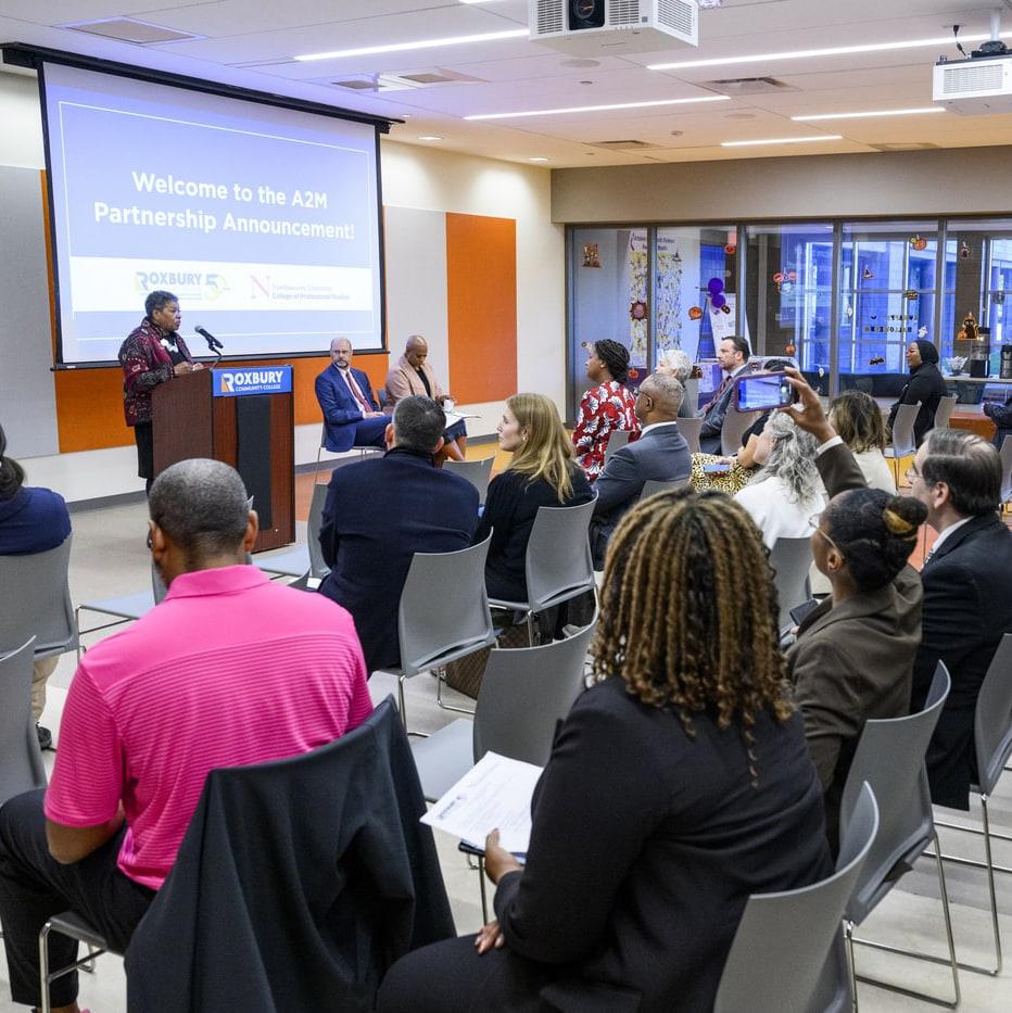 A woman speaks at a podium in a conference room with seated attendees. A screen behind her displays “Welcome to the A2M Partnership Announcement!” Several people sit at a panel table, and the room has a professional, collaborative atmosphere.