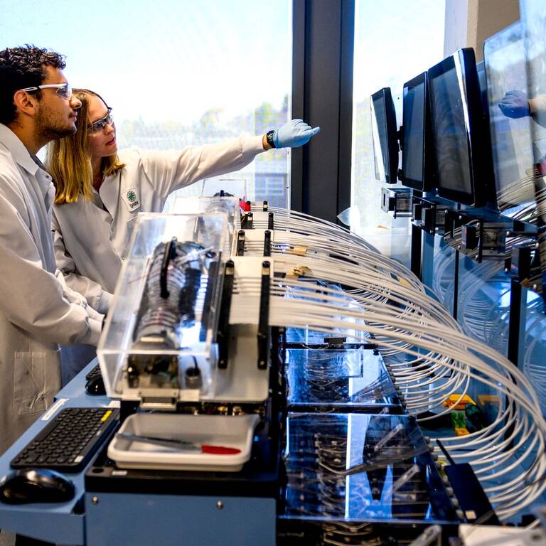 Two scientists in lab coats work at a laboratory station with multiple monitors, tubes, and scientific equipment. One scientist points at a screen while the other observes, both wearing safety glasses.