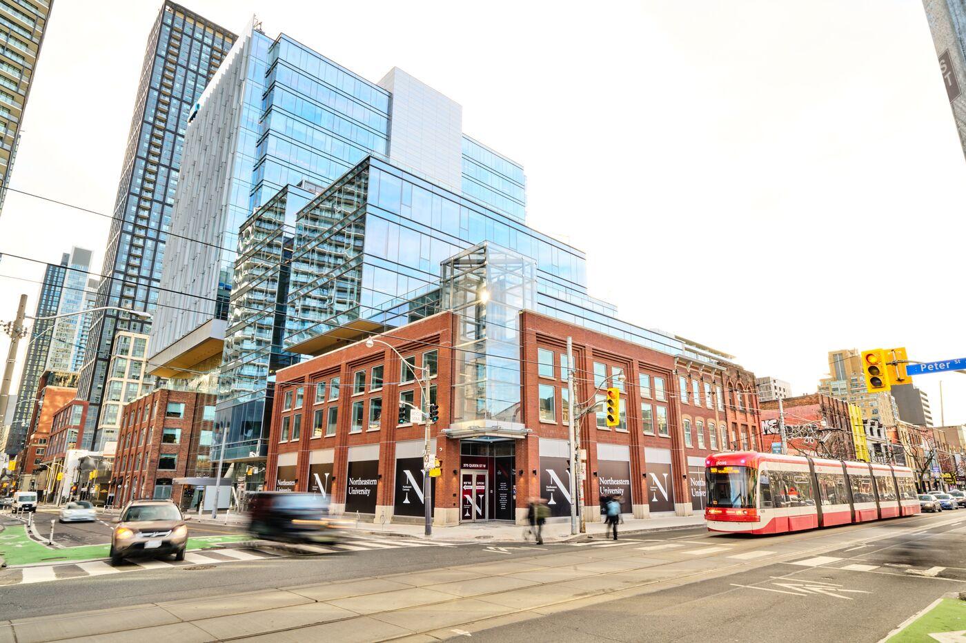 A modern glass building rises above a historic red-brick structure at a busy city intersection. A streetcar and several cars are stopped at a traffic light, with tall skyscrapers in the background under a bright sky.