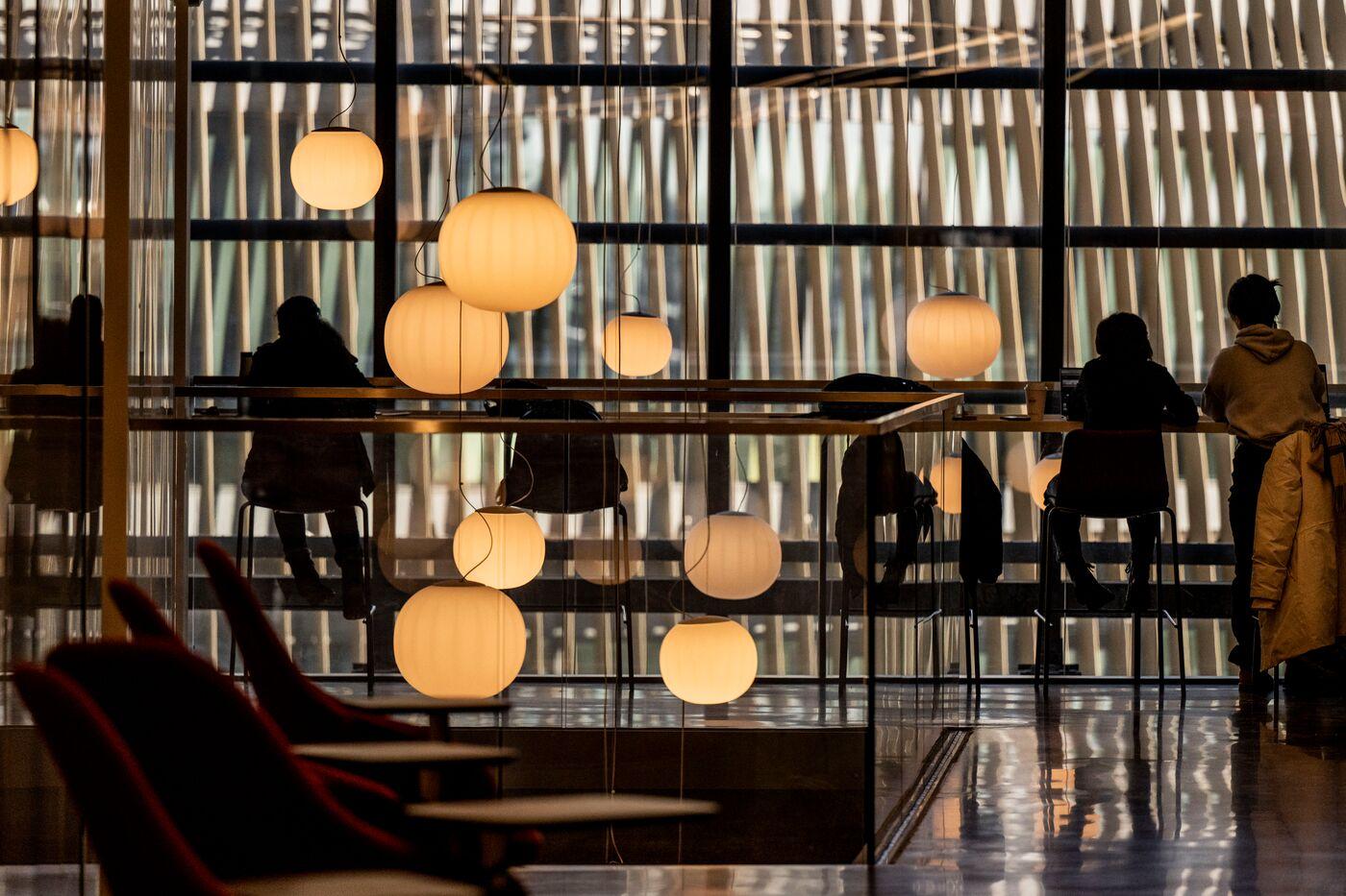 Silhouetted people sit at a long counter by a large window, with round hanging lights glowing warmly. Vertical lines on the window create an abstract, patterned backdrop. A coat hangs on a nearby chair.