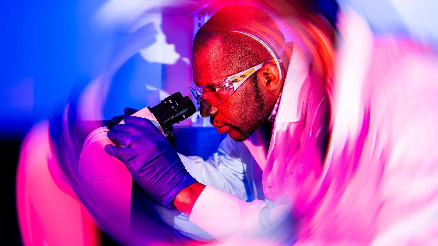 A scientist wearing safety glasses, gloves, and a lab coat looks into a microscope. The image is surrounded by a vivid, swirling pink and purple light effect, creating a dynamic, futuristic atmosphere.