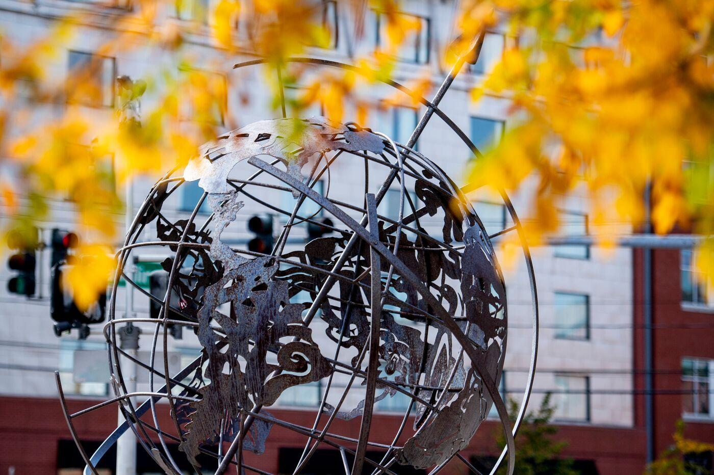 A metal globe sculpture with intricate designs stands outdoors, partially framed by yellow autumn leaves. In the background, there are traffic lights and a modern building with windows.