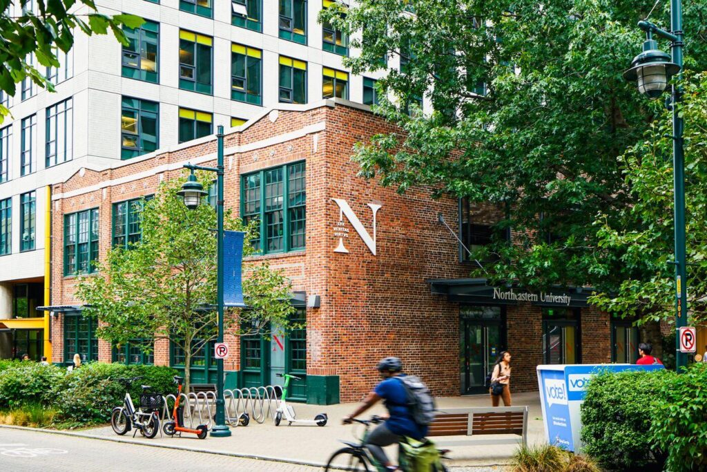 A brick building with a large letter "N" and sign for Northeastern University, surrounded by trees. A person rides a bike in the foreground, and another walks near the entrance. Scooters and bike racks are visible outside.