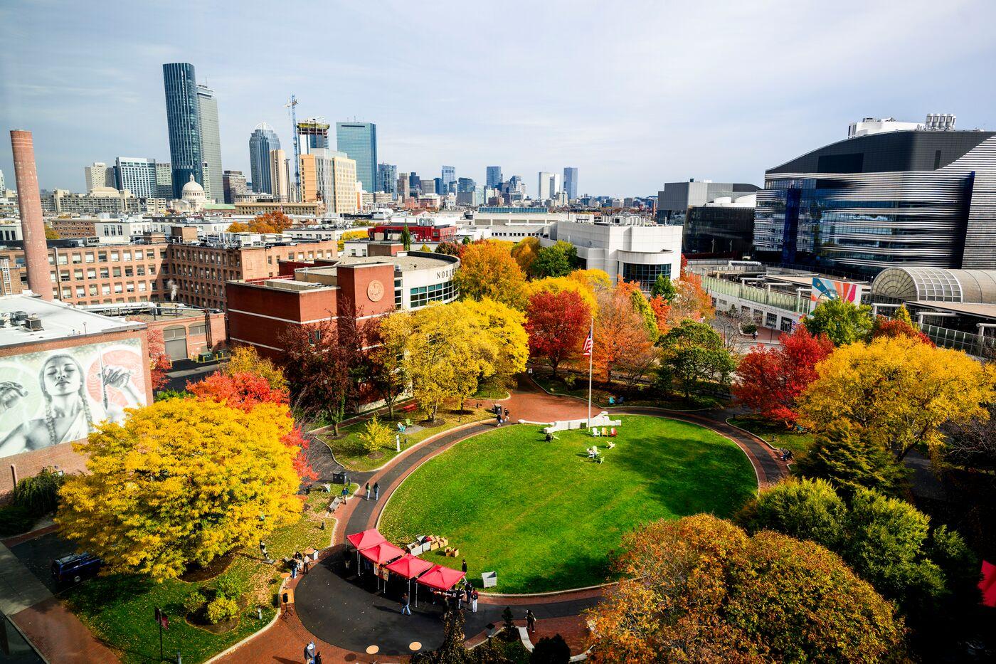 A cityscape view of a university campus with colorful autumn trees surrounding a green lawn, students walking and relaxing, and tall buildings in the background under a partly cloudy sky.