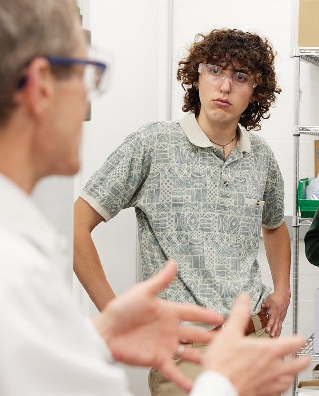A young person with curly hair and safety glasses listens attentively to someone speaking in the foreground, who is gesturing with their hands. They are in a room with white walls and metal shelves.