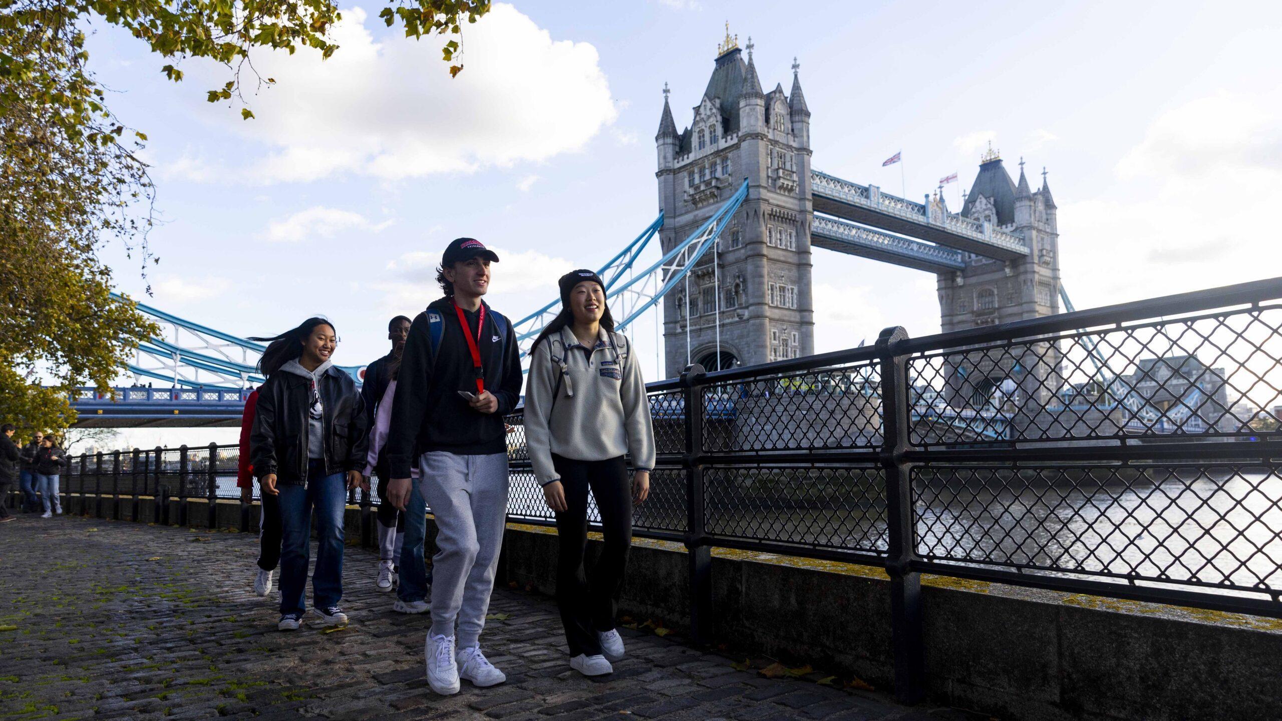 A group of young people walk along a riverside path near the iconic Tower Bridge in London on a sunny day, with trees and the river visible beside them.