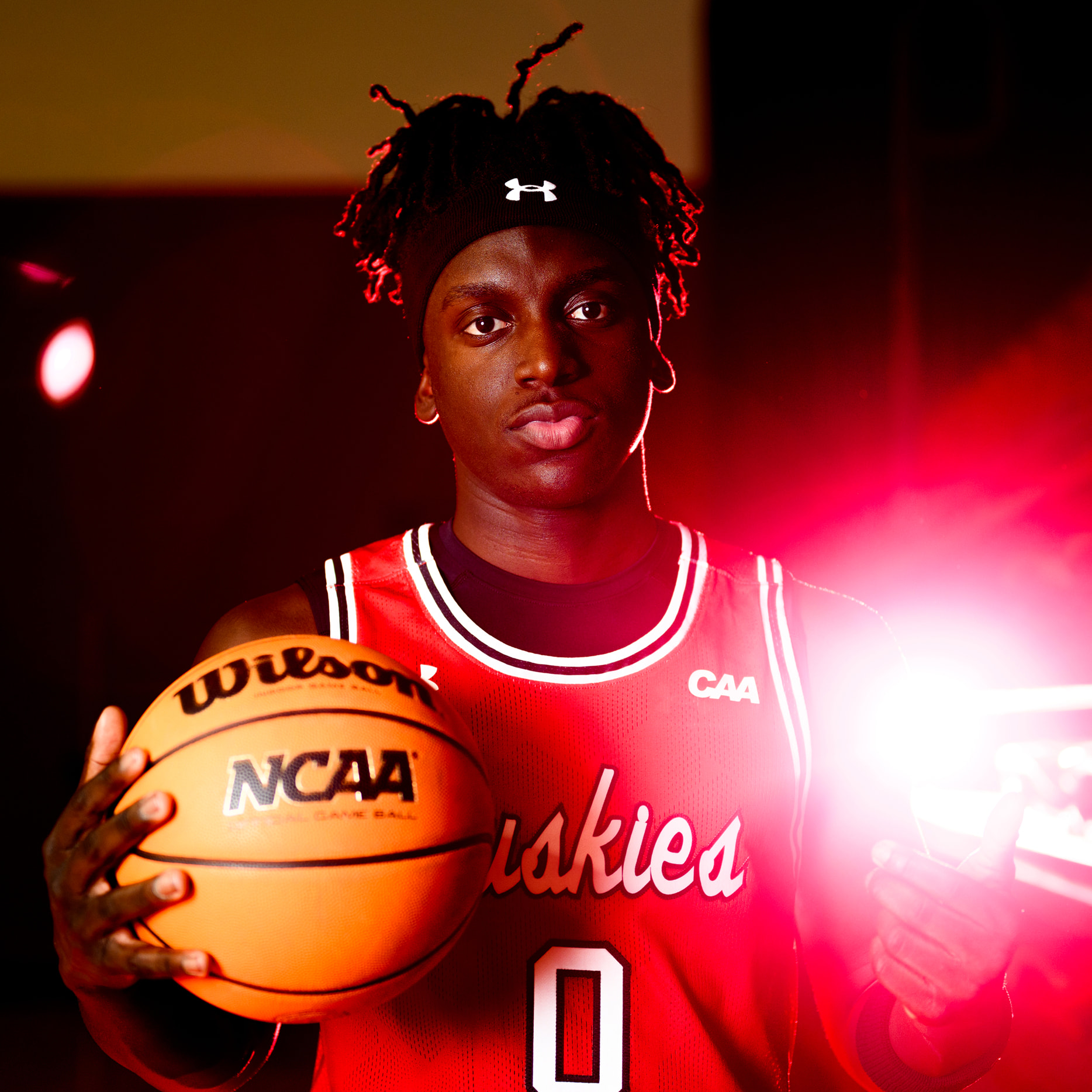 A basketball player in a red Huskies jersey with the number 0 holds a Wilson NCAA basketball. He stands in dramatic lighting with a bright red light flare behind him, looking directly at the camera.