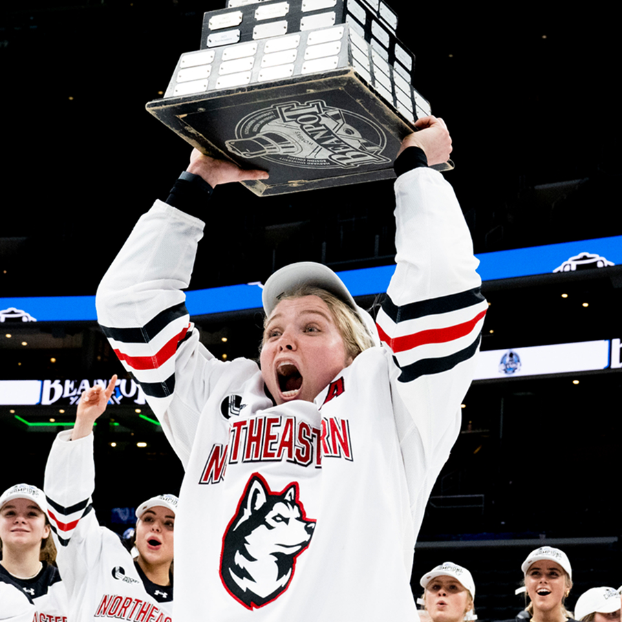 A Northeastern hockey player in a white jersey and cap excitedly lifts a large championship trophy overhead, surrounded by cheering teammates on an ice rink.