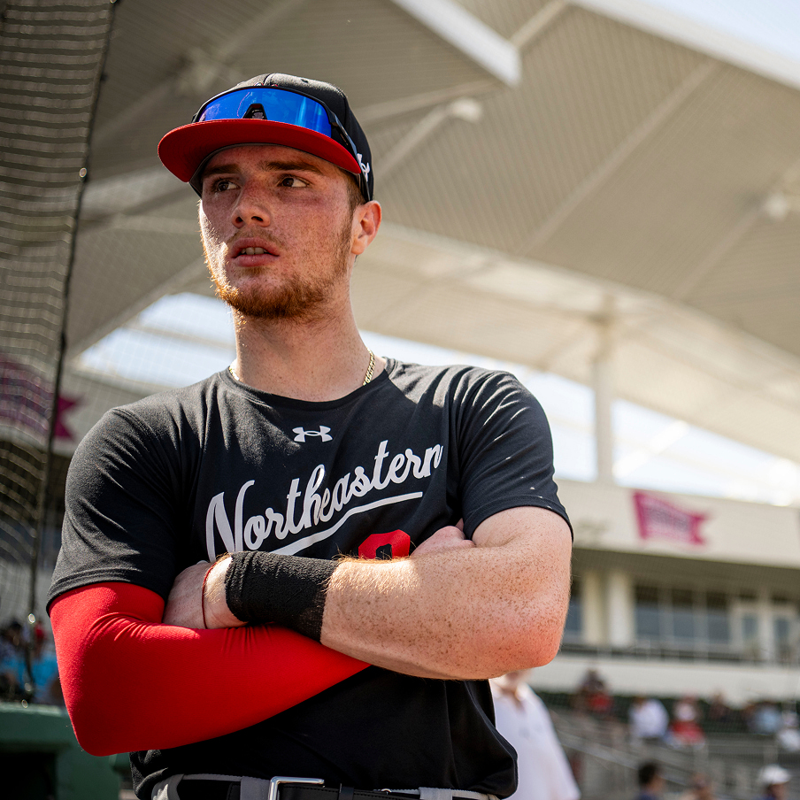 A baseball player in a black "Northeastern" jersey and red cap stands with arms crossed on a sunny field, wearing sunglasses on his cap and a red sleeve on his right arm. Stadium seating and a white roof are visible behind him.