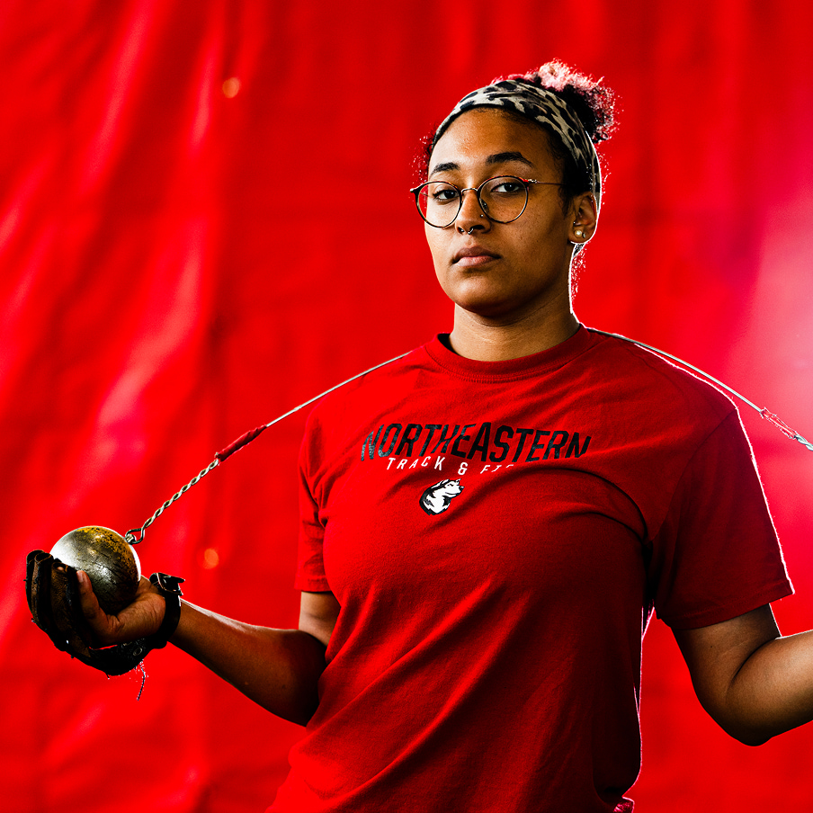 A person wearing glasses and a red "Northeastern Track & Field" shirt holds a metal ball with a chain, preparing for a hammer throw event. A bright red backdrop fills the background.