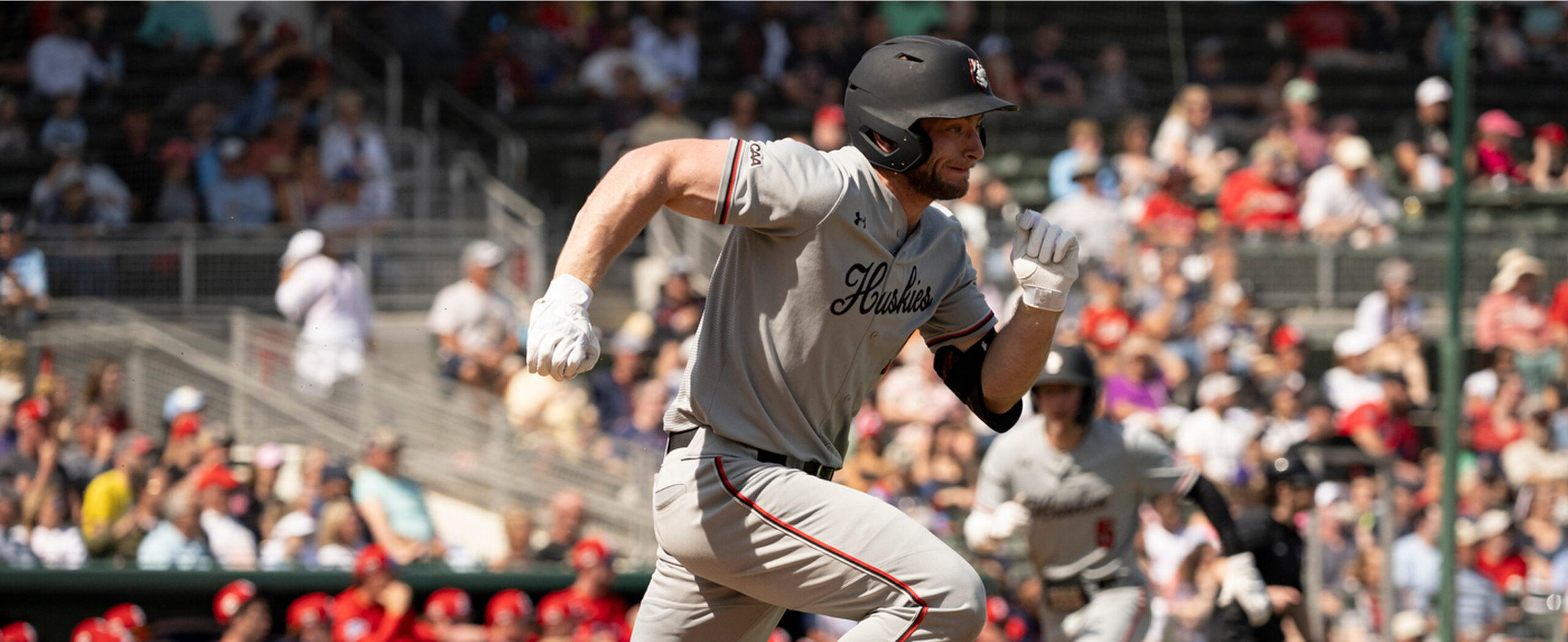 A baseball player in a gray uniform and black helmet sprints toward a base during a game, with a blurred crowd watching in the background.