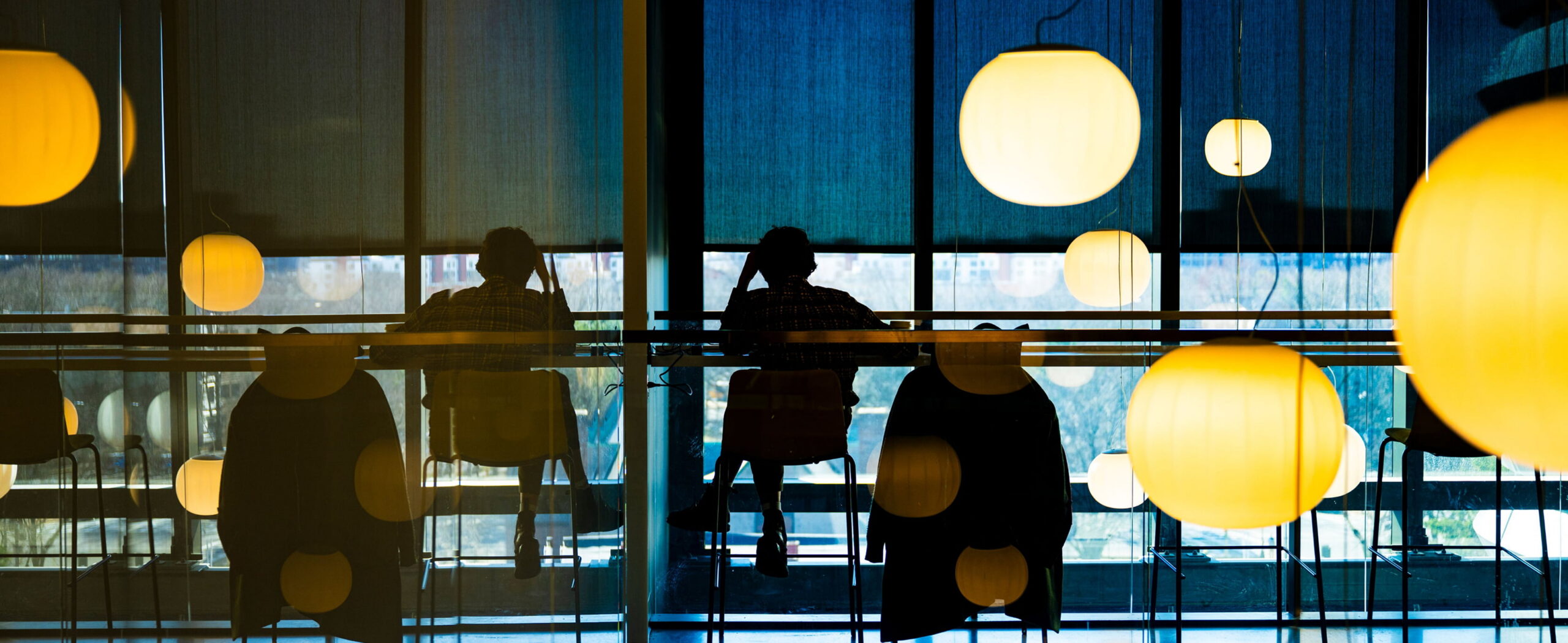 Silhouetted people sit at a long counter by a large window, with round hanging lights glowing warmly. Vertical lines on the window create an abstract, patterned backdrop. A coat hangs on a nearby chair.