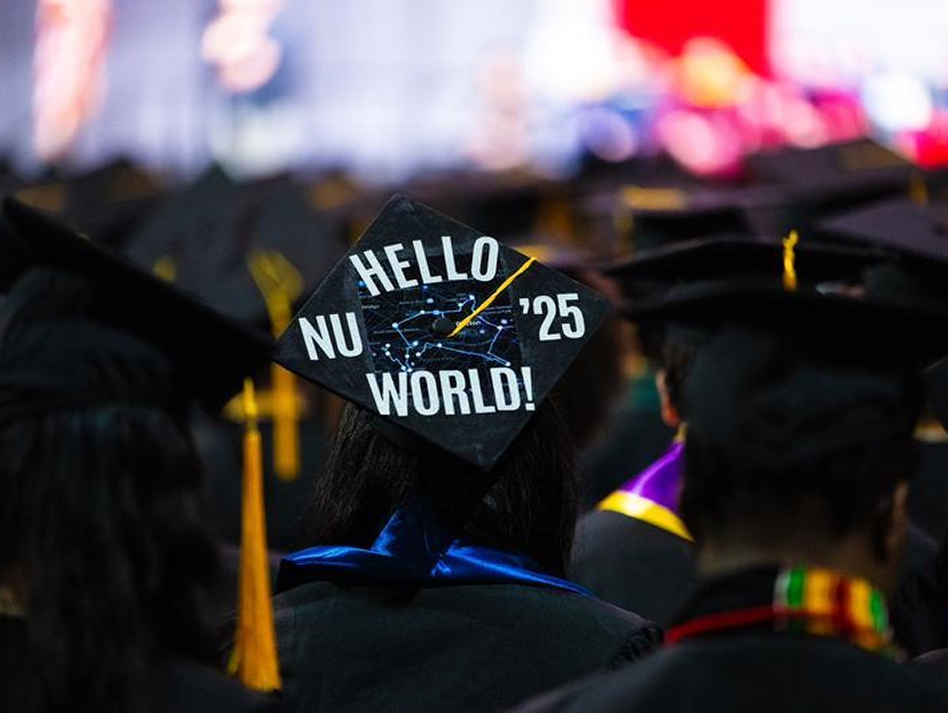 A graduate wears a decorated cap that reads "Hello World! NU '25" at a graduation ceremony, surrounded by other graduates in caps and gowns.