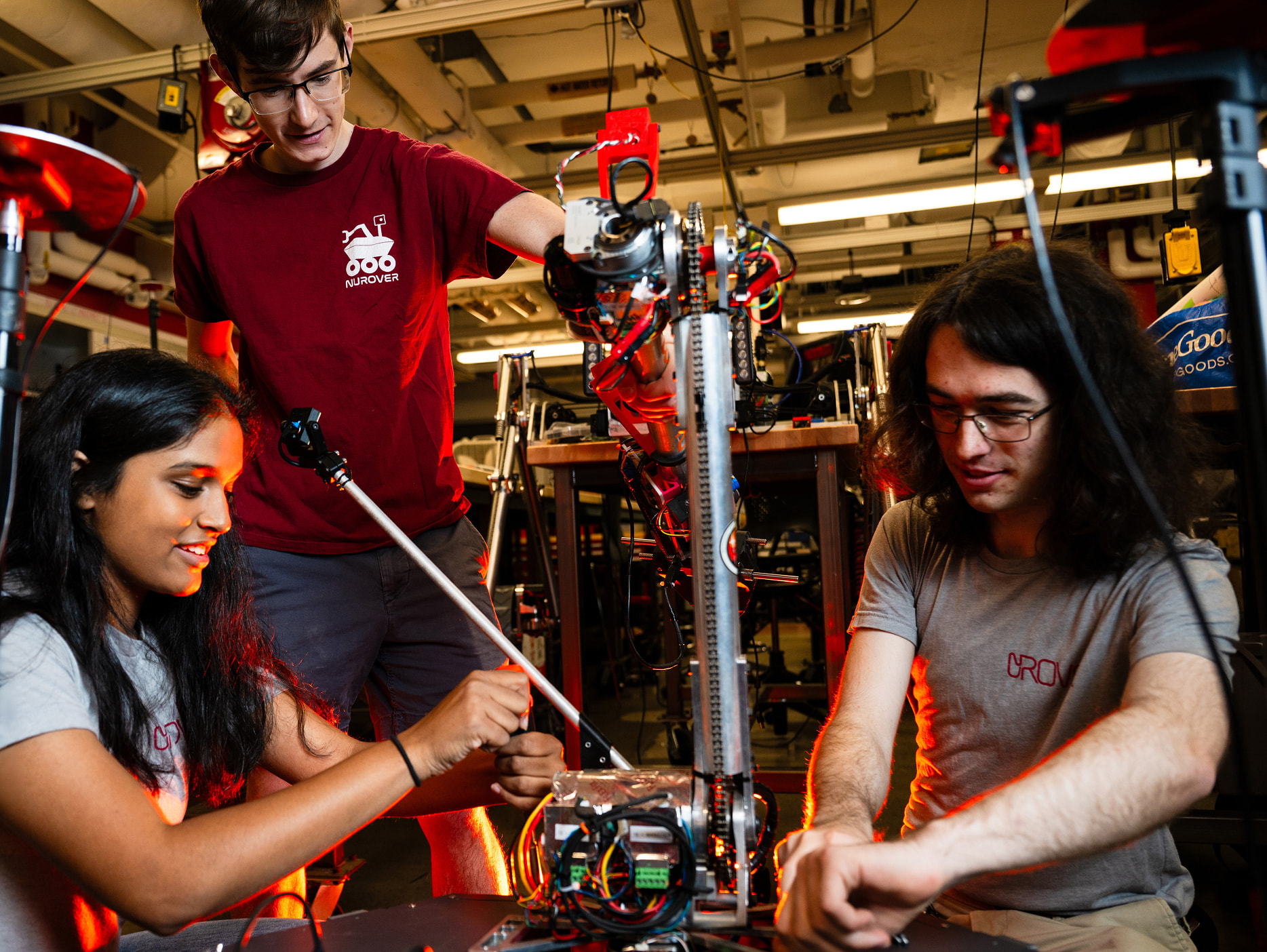 Three students work together on assembling or adjusting a robotic arm in a lab filled with technology and equipment, focused and engaged in their task.