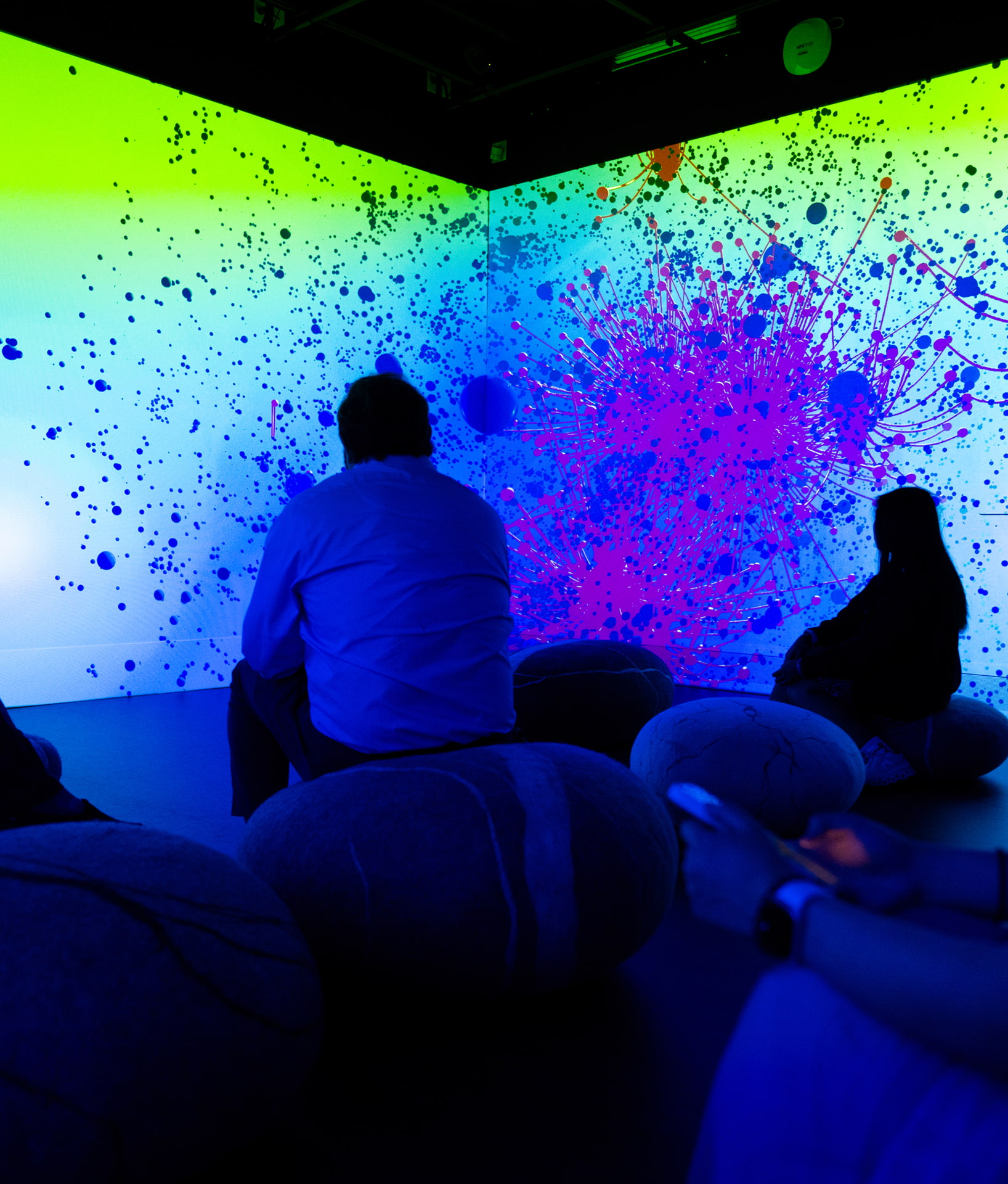 People sit on round cushions in a dark room, watching a vivid digital projection of blue, pink, and purple paint splatters on a green and yellow-lit wall. Some silhouettes are clearly visible in the foreground.