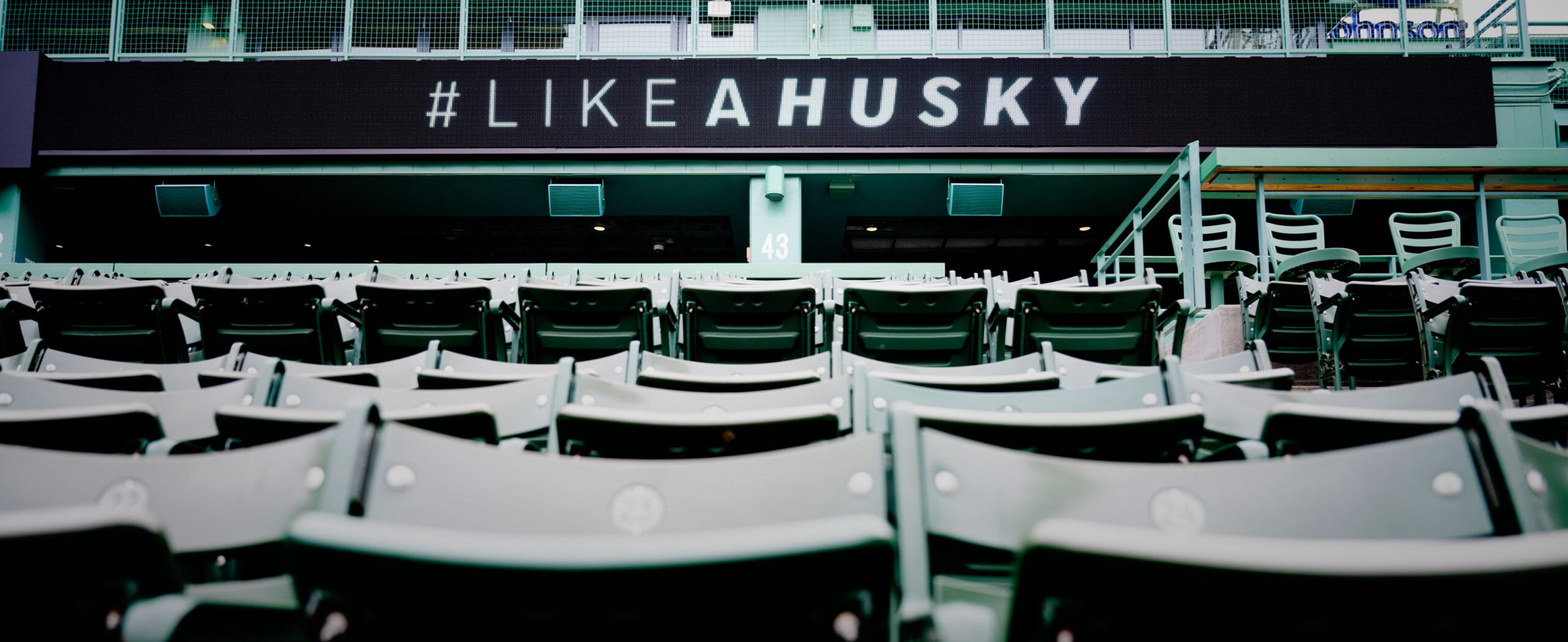 Empty stadium seats are shown in the foreground, with a large digital sign above displaying the text "#LIKEAHUSKY" in bold white letters.