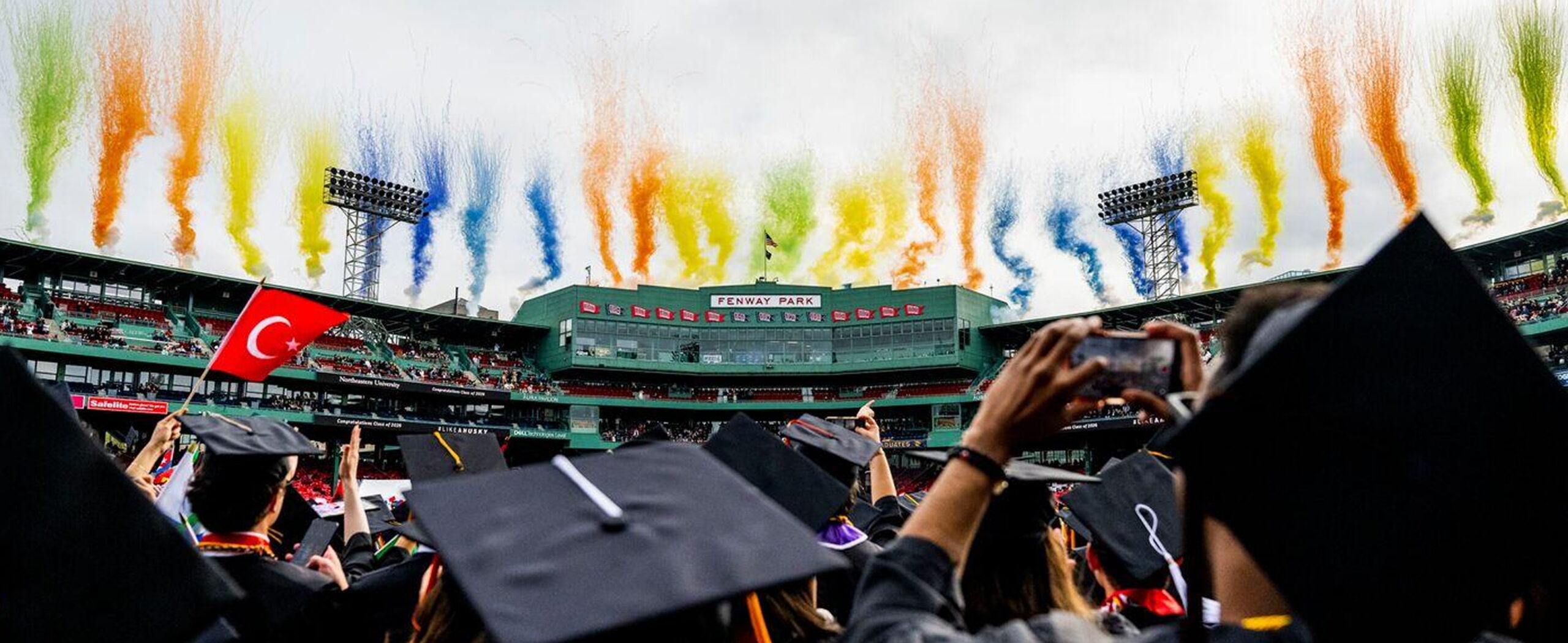 Graduates in caps and gowns face a stadium stage with colorful smoke and confetti rising into the sky; one person holds a Turkish flag and another takes a photo with a smartphone.