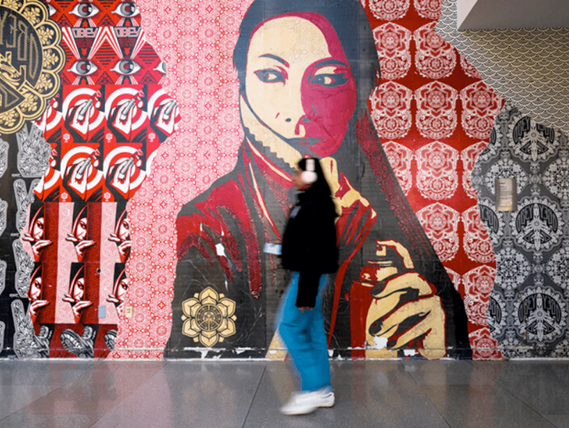A person in blue pants and a black jacket walks past a colorful mural featuring a large, stylized portrait of a woman with a patterned red and cream background and intricate designs.