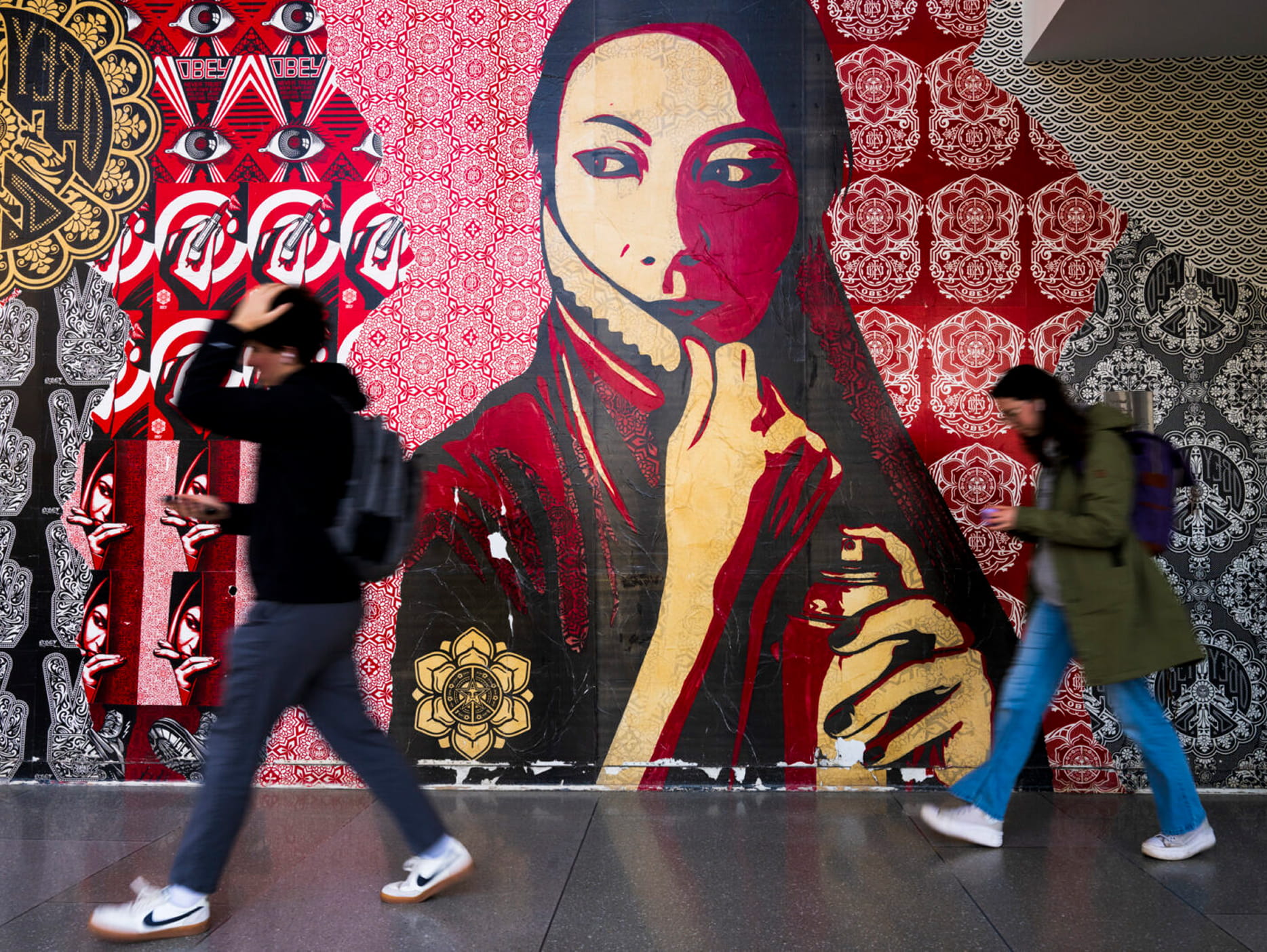 Two people walk past a colorful mural featuring a large, stylized portrait of a woman with a patterned red and cream background and intricate designs.