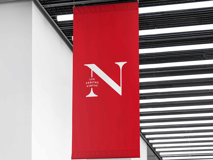 A red vertical banner displays a large white letter "N" with the Latin words "LUX VERITAS VIRTUS" in smaller white letters beside it, hanging indoors near a white wall and black ceiling slats.