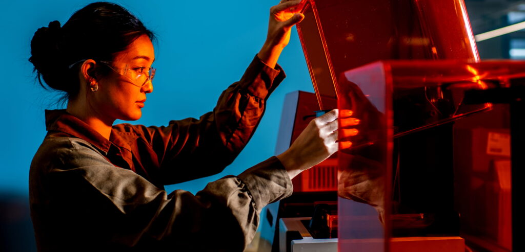 A person wearing safety glasses and a lab coat operates a 3D printer, lifting its orange-tinted cover in a well-lit, modern laboratory with a blue background.