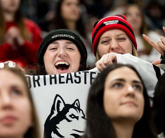 A group of excited Northeastern University fans, wearing team jerseys and sweatshirts, cheer and clap enthusiastically at a hockey game. One fan has "NU" painted on their face. The crowd is lively and energetic.