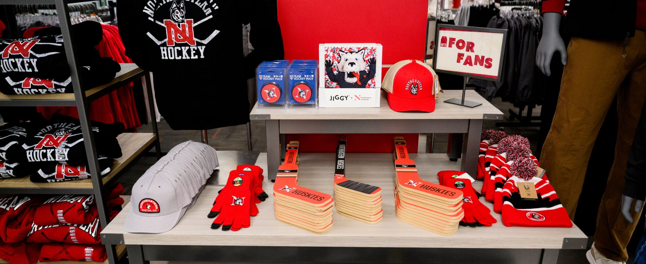 A store display features Northeastern University Huskies hockey apparel, including hats, gloves, scarves, shirts, and foam hockey stick clappers, arranged neatly on shelves with a "For Fans" sign.