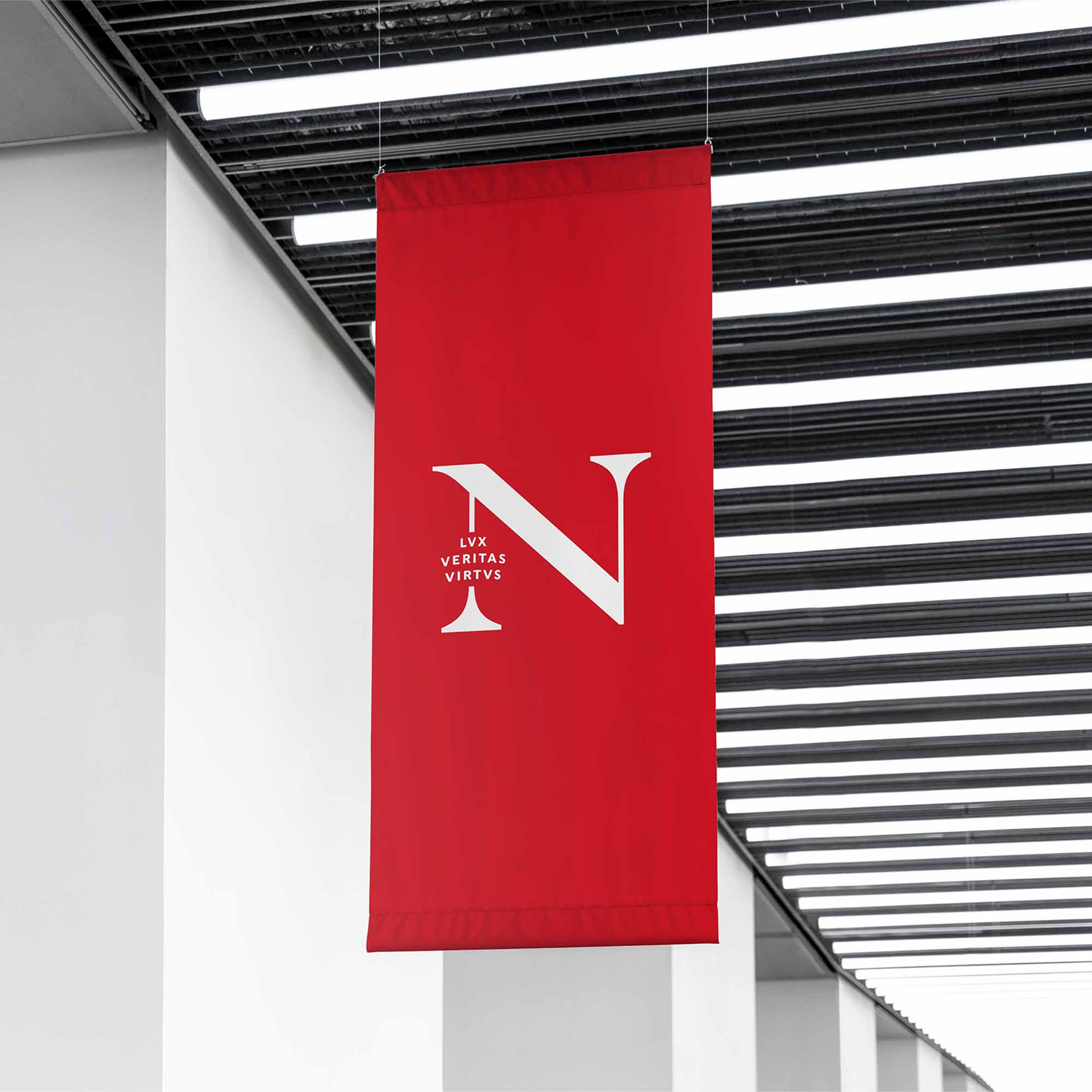 A red vertical banner displays a large white letter "N" with the Latin words "LUX VERITAS VIRTUS" in smaller white letters beside it, hanging indoors near a white wall and black ceiling slats.
