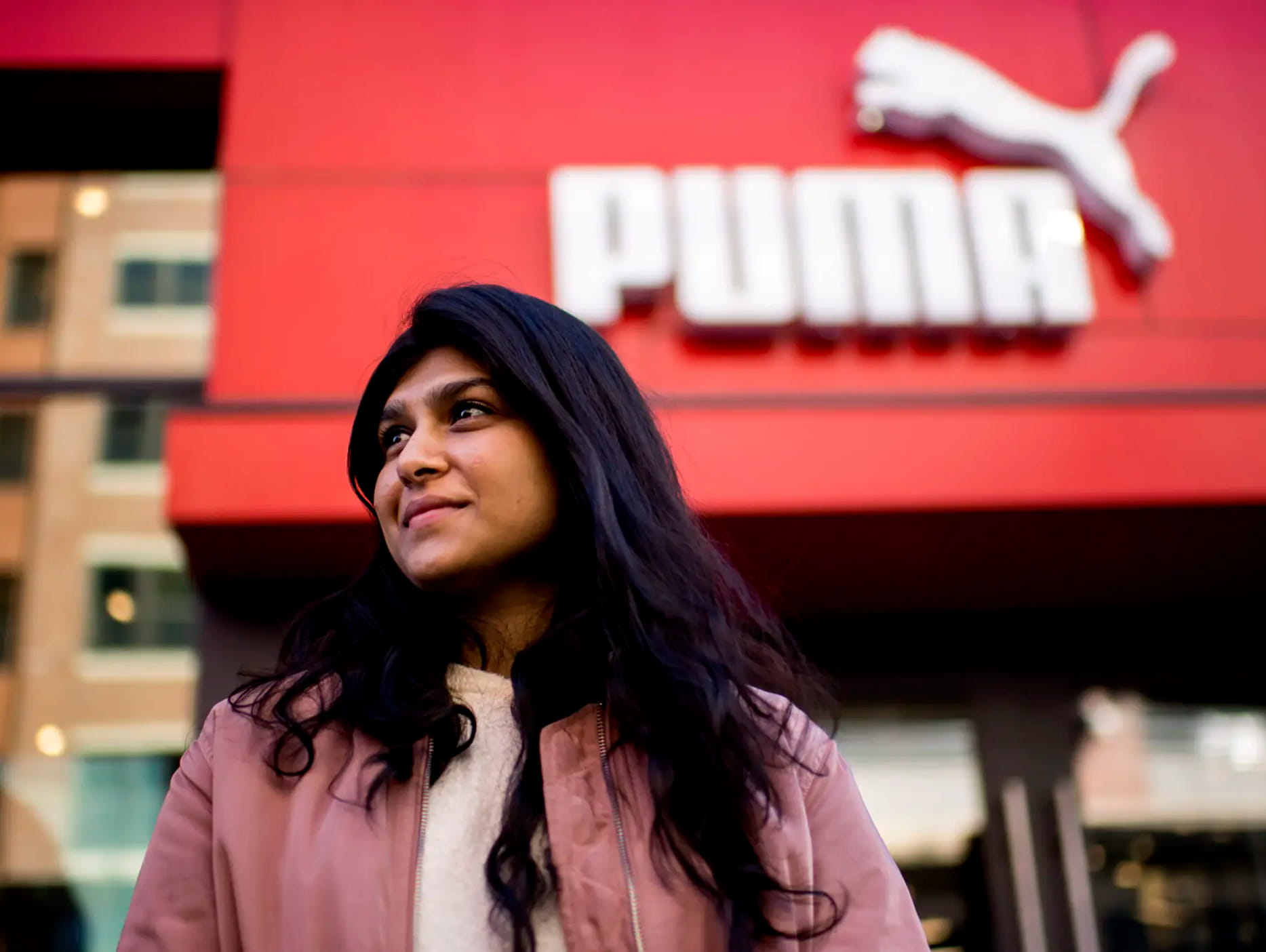 A woman with long dark hair stands in front of a Puma store with a red sign and white logo, looking off to the side and smiling slightly.