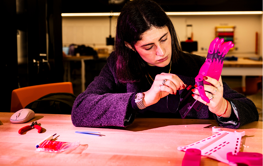 A woman sits at a workbench assembling a pink robotic hand, focused on attaching small components with tools and materials spread out on the table.