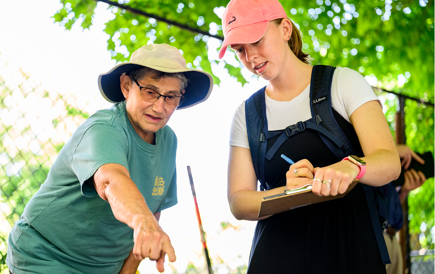 An older adult wearing a wide-brim hat points at something while speaking to a younger woman in a pink cap who is writing on a clipboard outdoors near a fence, with leafy trees in the background.