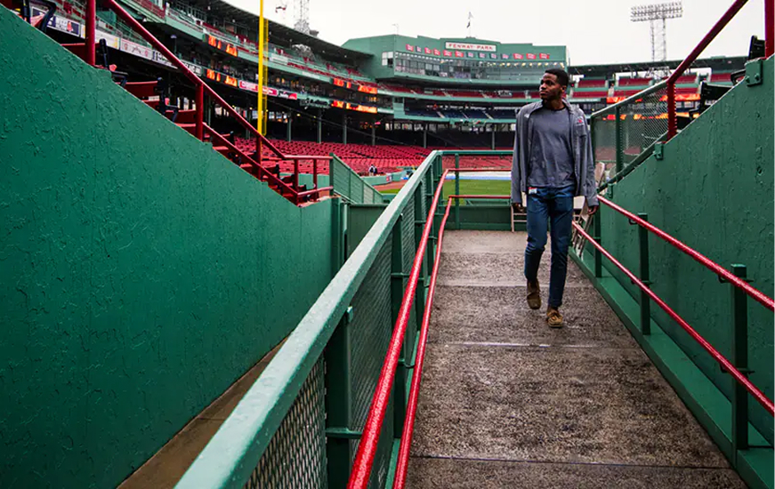 A man walks up a concrete ramp with red railings at Fenway Park baseball stadium, with empty green and red stands visible in the background on an overcast day.