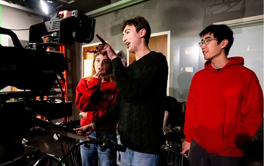 Three people stand next to a large video camera in a studio. One person points at the camera screen while explaining something to the others, who listen attentively. The group appears engaged in a discussion.