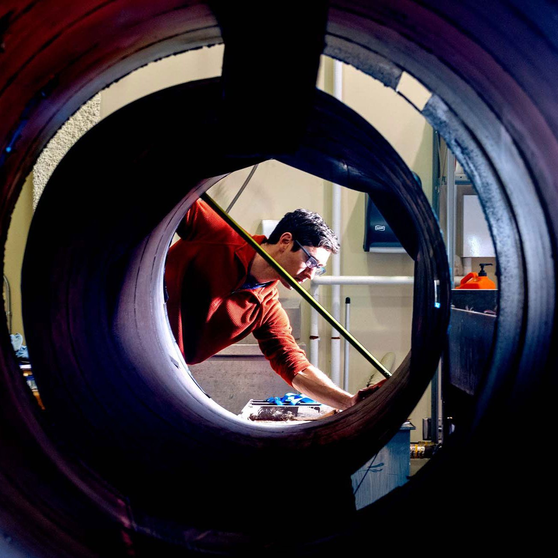 A person wearing safety glasses measures inside a large industrial pipe, seen through the circular opening, in a workshop setting with tools and equipment around.