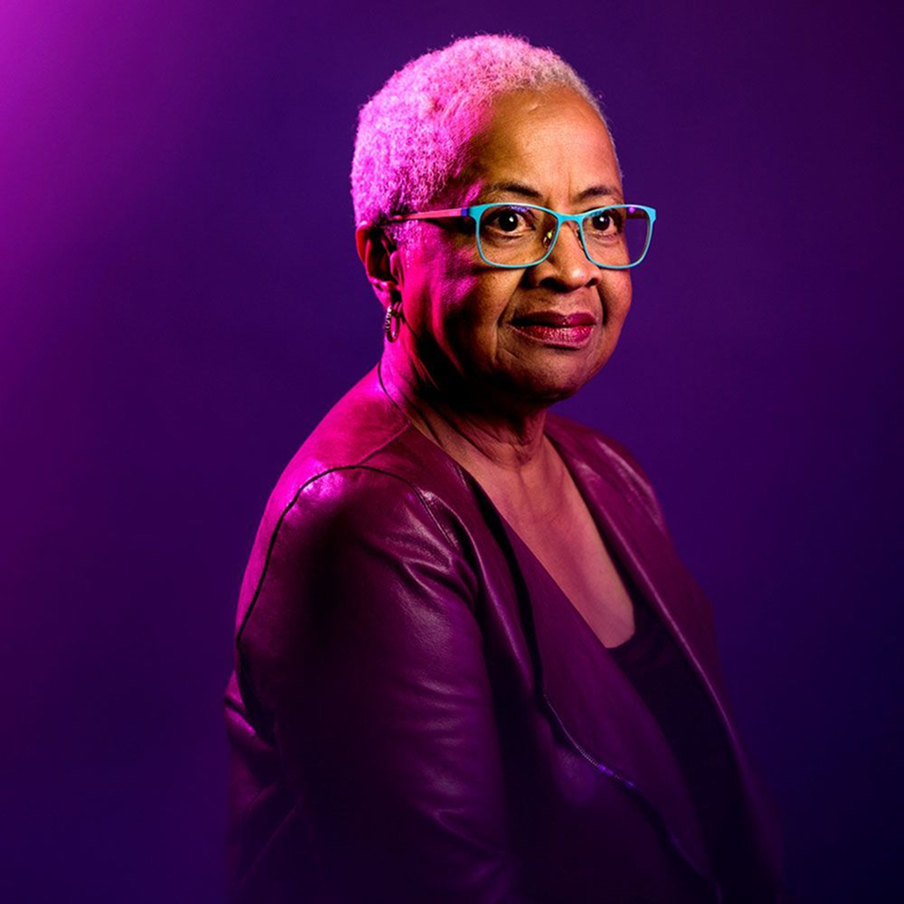 A woman with short hair and teal glasses wears a burgundy jacket and smiles softly against a pink and purple gradient background.