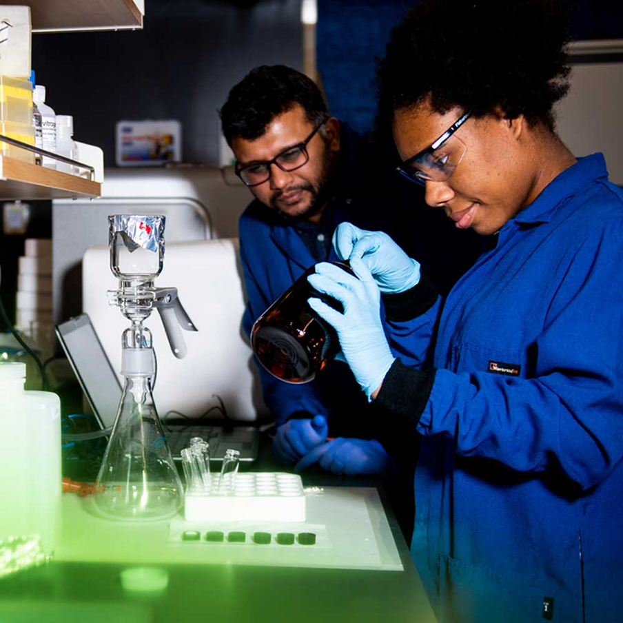 Two scientists in blue lab coats and gloves work in a laboratory. One is pouring liquid from a dark bottle, while the other observes. Lab equipment and containers are visible on the bench.