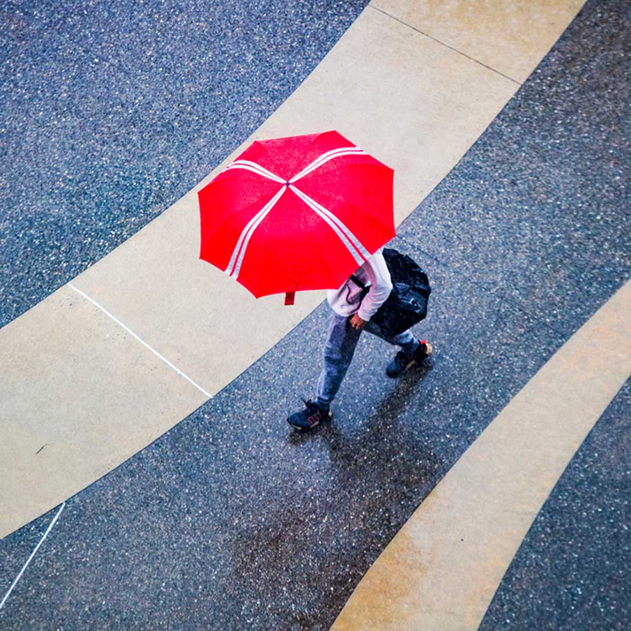 A person walking on a wet, curved pathway holds a bright red umbrella, seen from above. The ground has contrasting light and dark sections, and the person carries a black backpack.
