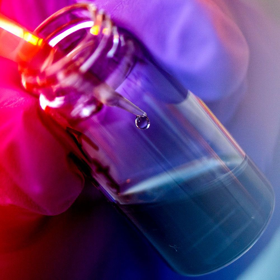 A close-up view of a gloved hand holding a small glass vial while a dropper releases a droplet of liquid inside, illuminated by vibrant purple and orange lighting.