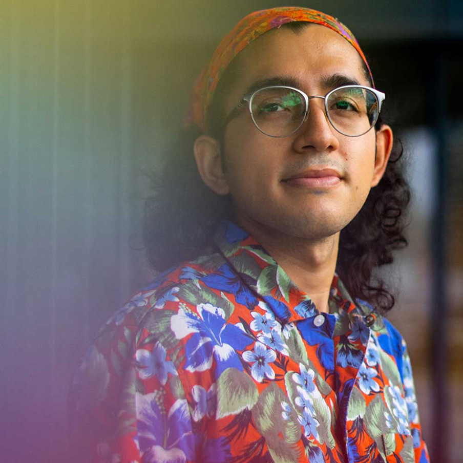 A person with long dark hair, glasses, and a colorful floral shirt looks at the camera. They wear a red headband and stand against a softly blurred background with warm and cool tones.