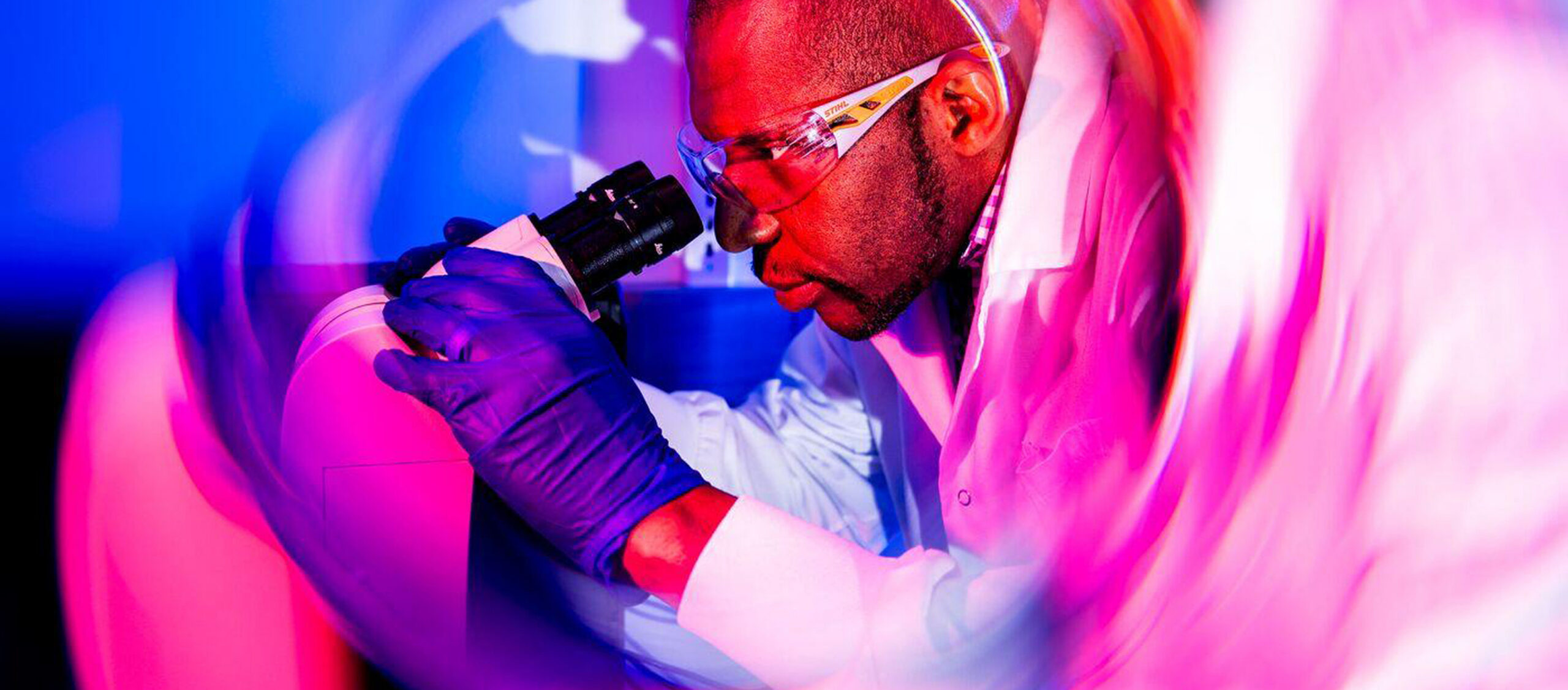 A scientist wearing safety glasses, gloves, and a lab coat looks into a microscope. The image is surrounded by a vivid, swirling pink and purple light effect, creating a dynamic, futuristic atmosphere.