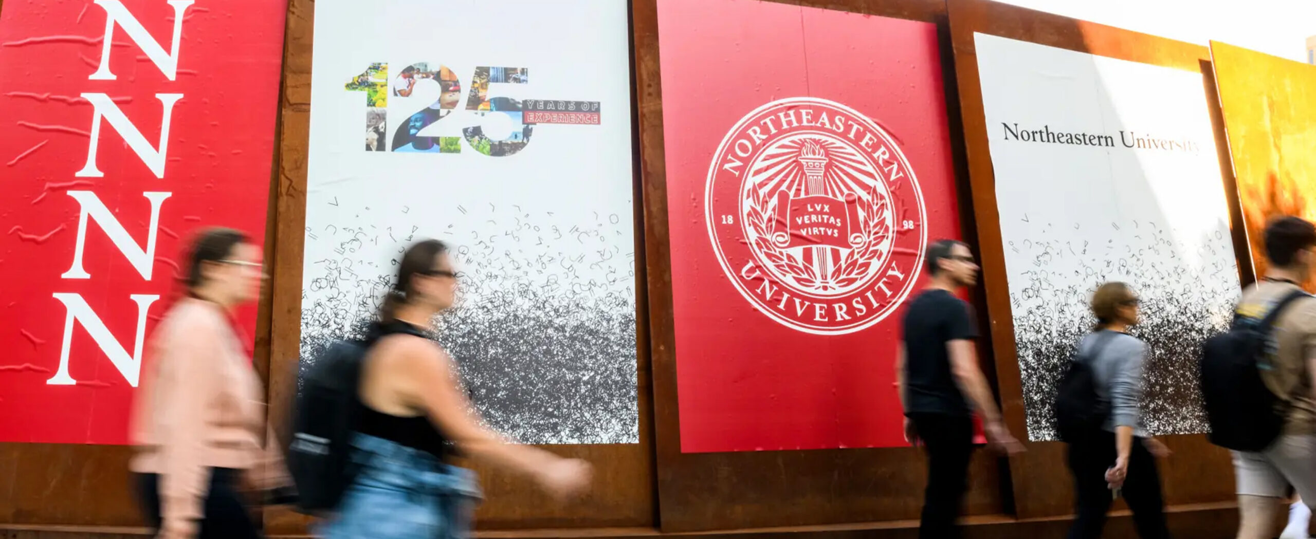 Students walking past a series of northeastern logos posted on a wall