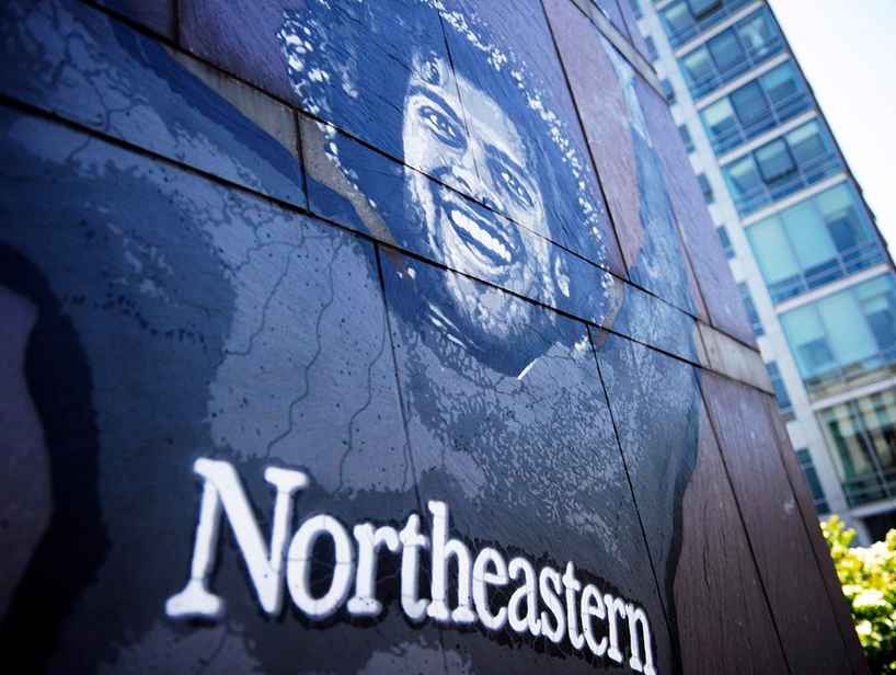 A mural featuring a smiling person with curly hair is painted on a wall, above the word "Northeastern." A modern glass building is visible in the background.