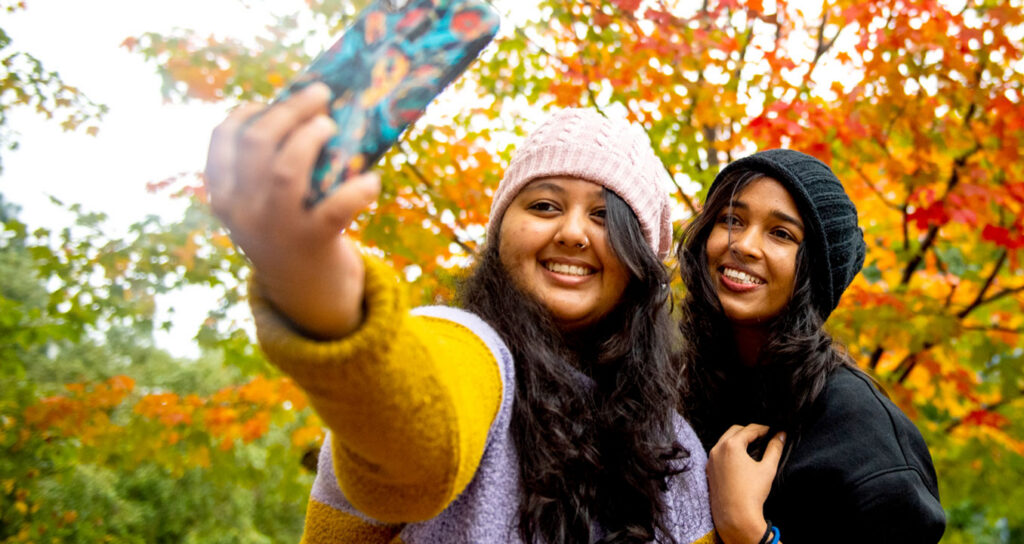 Two students pose for a selfie with fall foliage and Northeastern banners in the background