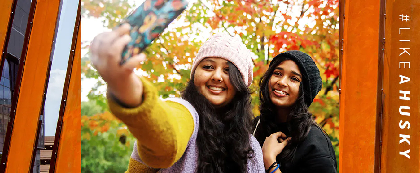 Two students pose for a selfie with fall foliage and Northeastern banners in the background