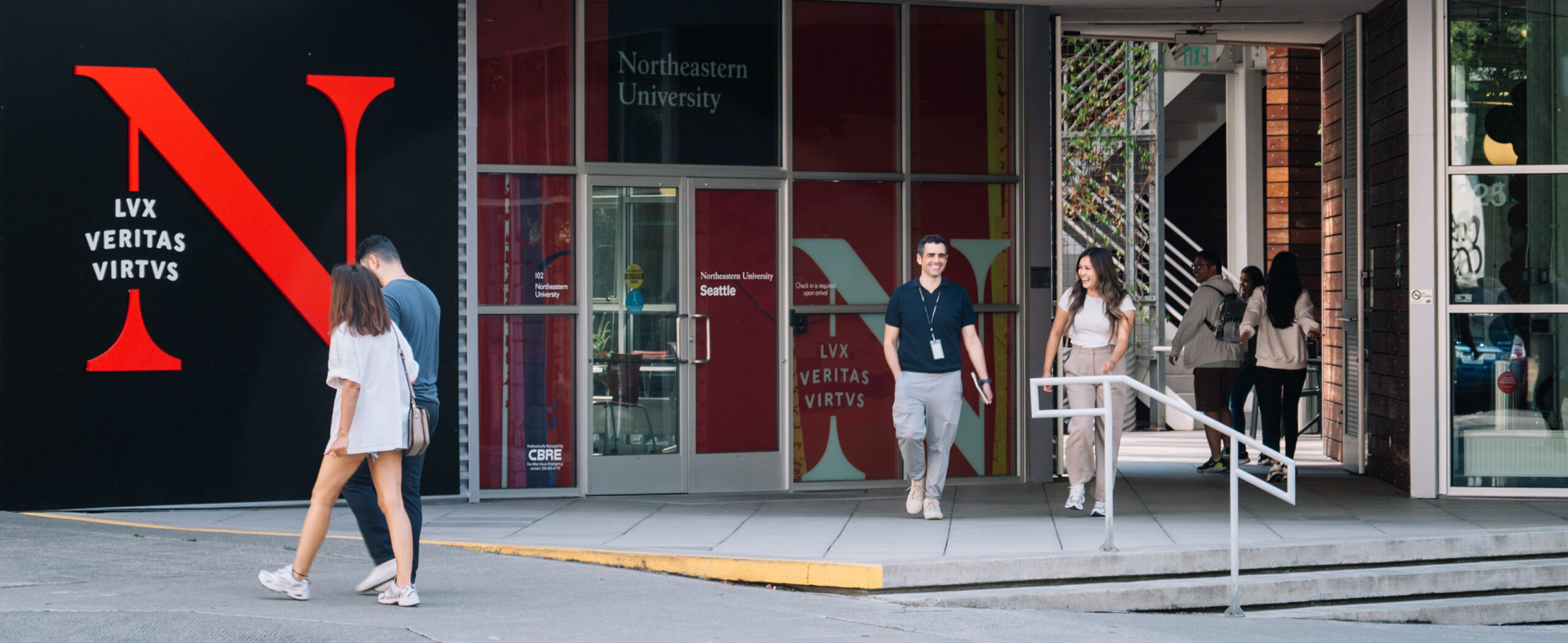 Exterior of Northeastern University's Seattle campus building, with a large red 'N' logo and the words 'LUX VERITAS VIRTUS' on a black wall to the left. Students and pedestrians walk along the sidewalk and near the entrance, where glass doors display 'Northeastern University Seattle' signage.